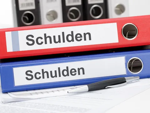 Ein Stift und zwei Aktenordner mit der Aufschrift 'Schulden' liegen vor einer Reihe weiterer Ordner
