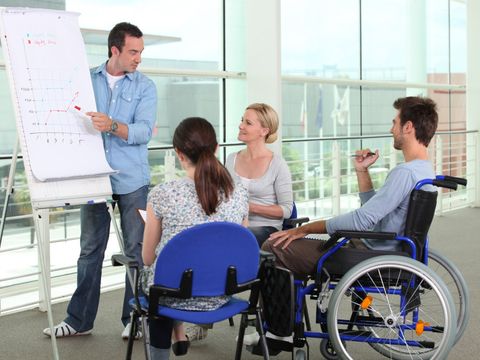 Besprechung unter 4 MItarbeitern, zwei sitzen auf einem Stuhl, einer im Rollstuhl und der vierte steht an einem Flipchart und erklärt ein Diagramm