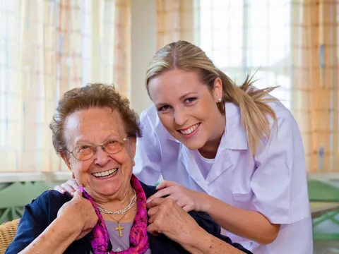 Eine Betreuerin lächelt mit einer Seniorin in die Kamera