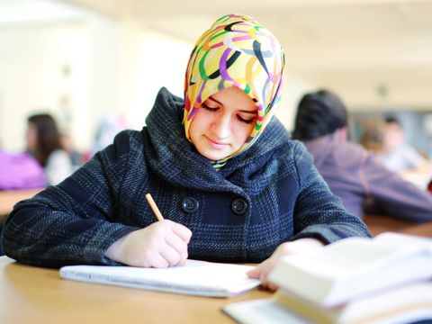Junge Muslima sitzt an einem Tisch und schreibt ewas in einen Block