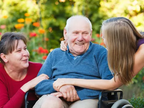 Senior im Rollstuhl wird von seiner Tochter und Enkelin umarmt. Alle drei lächeln sich an.