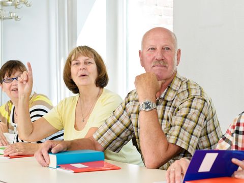 Senioren sitzen an einem Tisch mit Büchern, eine Seniorin hebt den Zeigefinger und setzt zum Sprechen an