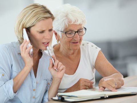 Seniorin zeigt auf eine Seite in einem Terminkalender, eine junge Frau sitzt daneben und telefoniert