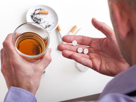 Mann mit Tabletten und Whiskyglas in den Händen und benutztem Aschenbecher im Hintergrund