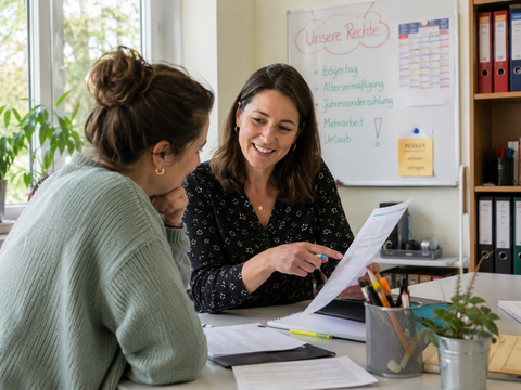 Zwei Frauen sitzen in einem hellen Büro an einem Tisch und besprechen gemeinsam Unterlagen. Eine Frau zeigt lächelnd auf ein Dokument. Im Hintergrund hängt ein Whiteboard mit Stichpunkten zu arbeitsrechtlichen Themen wie Urlaub, Mehrarbeit und Jahressonderzahlung.