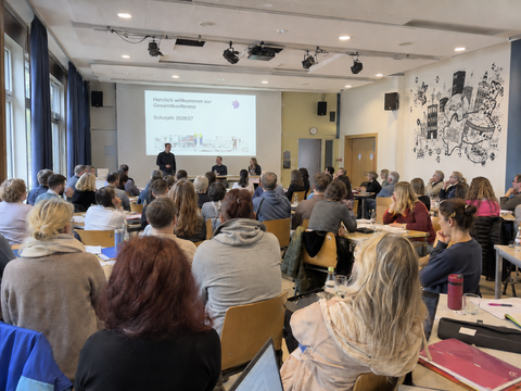 Ein großer Konferenzraum in einer Schule ist mit zahlreichen Lehrkräften und Beschäftigten besetzt, die an Tischen sitzen und nach vorne zur Präsentation schauen. Auf der Leinwand steht „Herzlich willkommen zur Gesamtkonferenz“ und darunter „Schuljahr 2026/27“. Vorne stehen und sitzen Mitglieder der Schulleitung.
