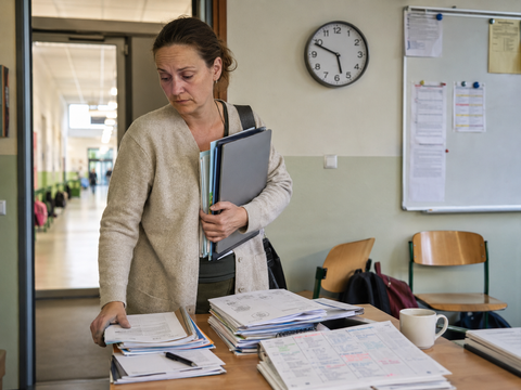 Eine erschöpft wirkende Lehrerin steht mit Laptop, Ordnern und Unterlagen am Schreibtisch. Im Hintergrund sind ein Schulflur, eine Uhr und eine Tafel mit Aushängen zu sehen. Die Szene vermittelt Arbeitsbelastung, Zeitdruck und zusätzliche Aufgaben im Schulalltag.