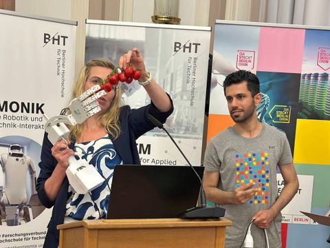 Zwei Vortragende halten eine Roboterhand, die Tomaten pflückt