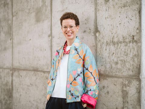 Eine Frau mit kurzen, lockigen Haaren und einer bunten Jacke steht vor einer Betonwand und lacht.