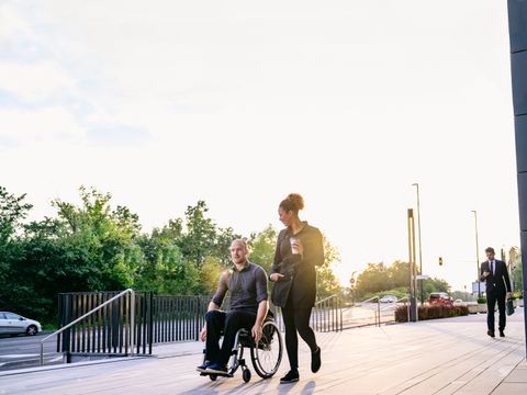Zwei Personen, davon eine im Rollstuh sitzend unterhalten sich draußen auf einem Gehweg.