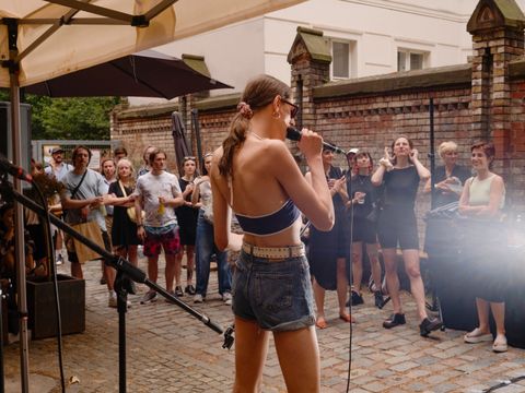 Eine sommerlich gekleidete Person mit langen Haaren singt vor einer Gruppe Menschen in einem Innenhof.