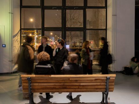 Menschen auf einer Parkbank, dahinter Menschen im Gespräch vor einem großen Fenster