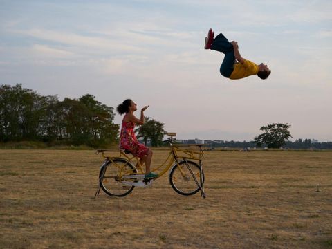 Eine Frau auf einem Fahrrad in einem Feld. Ein Akrobat macht einen Salto in der Luft.