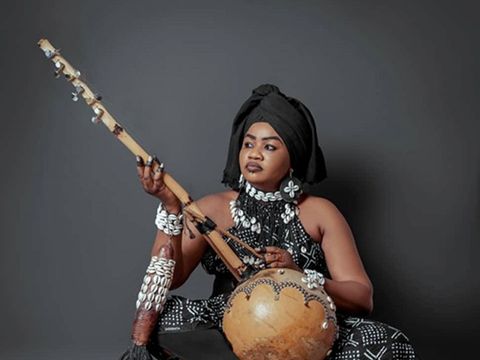 Eine Musikerin aus Burkina Faso im Schneidersitz mit einem Musikinstrumet vor grauem Hintergrund