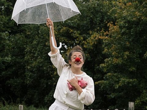 Eine in weiß gekleidete fröhliche Frau auf einer Wiese mit Koffer, Regenschirm und Clownsnase