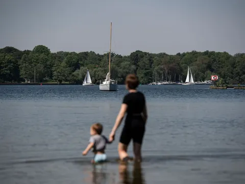 Person mit Kind watet am Seeufer, im Hintergrund Segelboote auf ruhigem Gewässer.