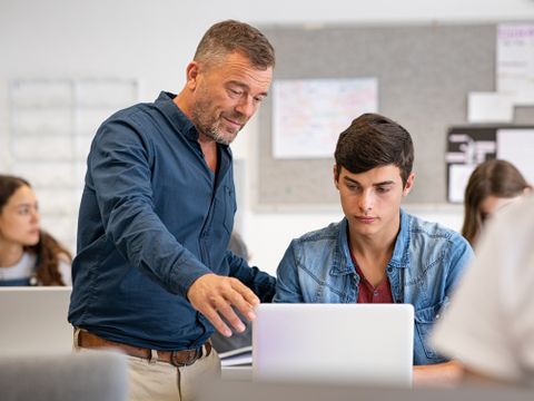 Medienpädagogik - Lehrer steht bei einem Schüler am Laptop im Klassenzimmer