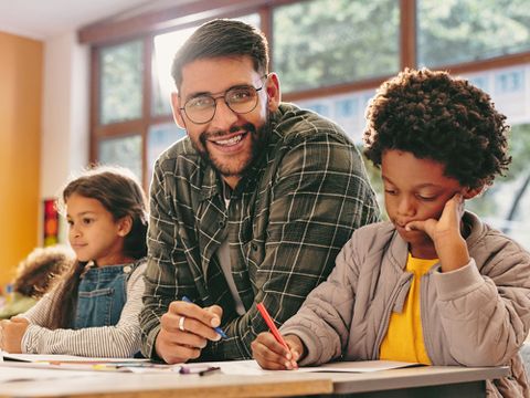 Pädagogische Unterrichtshilfe - Klassenzimmer mit Kindern und einem lächelnden Mann neben zwei Kindern