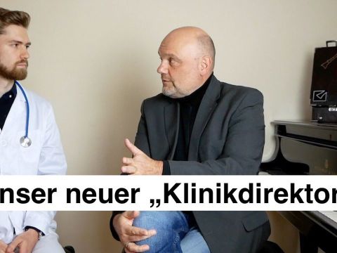 Musikschuldoktor im weißen Kittel sitzt neben Musikschulleiter im grauen Jackett