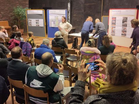 Menschen sitzen in einem Raum vor Stelltafeln