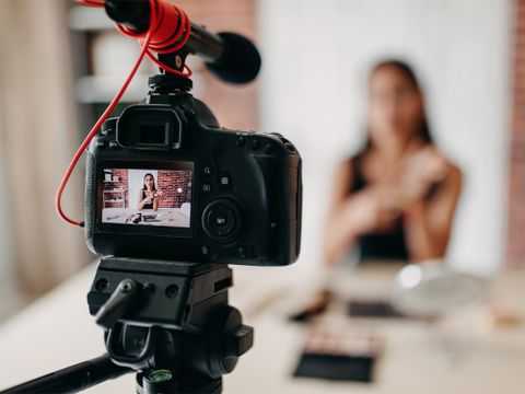 Im Vodergrund eine Kamera auf einem Stativ aufgebaut, welche eine Frau bei einem Video filmt