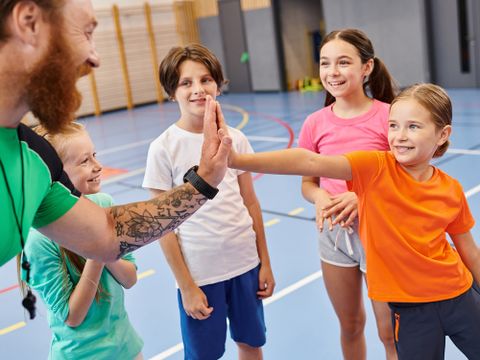 Gruppe junger Kinder steht begeistert in einer Turnhalle und klatschen mit dem Lehrer ein