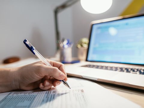 Person arbeitet an Schreibtisch und füllt ein Fomular aus, ein Laptop steht dahinter
