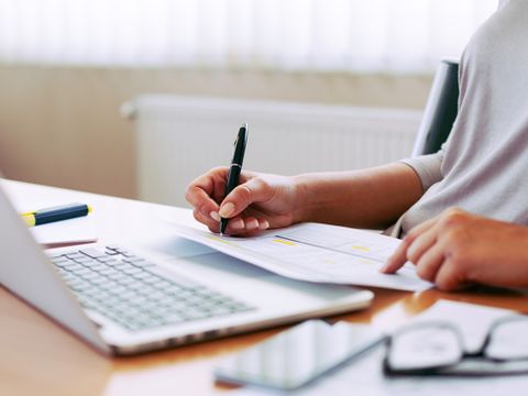 Frau sitzt an Arbeitsplatz und arbeitet sowohl mit einem Laptop als auch mit Papierunterlagen