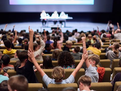 Viele Kinder im Saal bei einer Vorlesung. EInige melden sich