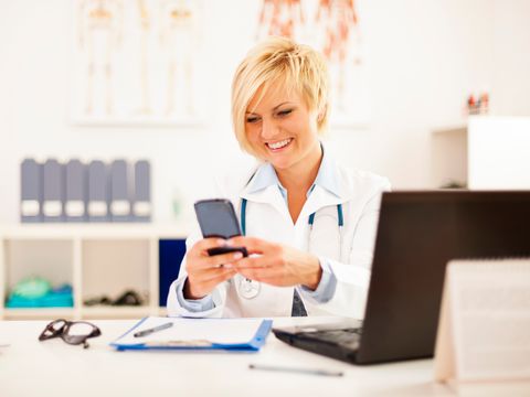 Eine Frau sitzt an einem Schreibtisch in ihrem Sprechzimmer einer Arztpraxis. Vor ihr sieht man die Umrisse des Bildschirms eines Computers. Sie schaut auf das Handy, das sie in ihren Händen vor sich hält. Dabei lächelt sie. Sie trägt einen weißen Arztkittel und ein Stethoskop um den Hals.