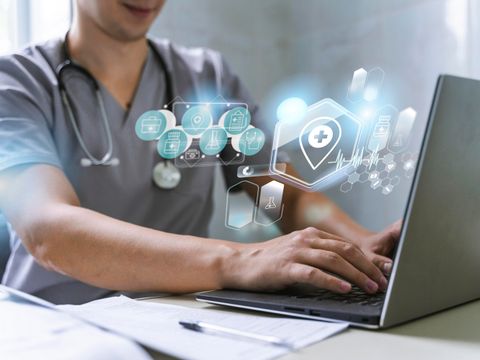 Eine Person mit einem Stethoskop um den Hals und einem Arztkittel sitzt an einem Laptop und tippt auf der Tastatur. Neben dem Laptop liegen Papiere und ein Stift. Aus dem Laptop Bildschirm heraus kommen blau und weiß leuchtende Symbole aus dem Bereich Medizin, z.B. ein Erste-Hilfe-Kreuz.