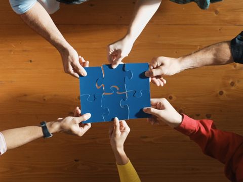 Sechs Unterarme sind in der Draufsicht zu sehen. Die zugehörigen Hände halten jeweils ein blaues Puzzleteil. Sie fügen die Puzzleteile in der Mitte so zusammen, dass sie ineinanderpassen.