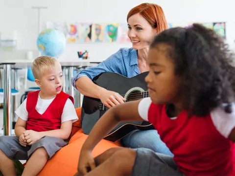 Schulkinder und Lehrerin mit Gitarre sitzen im Klassenzimmer, ein Kind mit geistiger Behindung