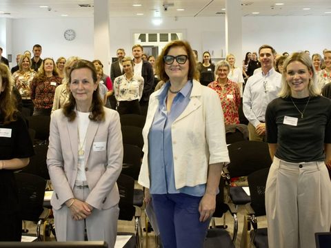 Pressefoto der Bündnistagung mit den Akteurinnen des Projekts "Fachkräftesicherung in der Pflege" und den Teilnehmenden. Vorne stehen v.l.n.r.: Katrin Mauch von ArbeitGestalten, XX, Senatorin Dr. Ina Czyborra, Leonie Teubel von der Senatsverwaltung für Wissenschaft, Gesundheit und Pflege und Sandra Tausch von ArbeitGestalten. Sie alle stehen in einem hellen Raum und blicken lächelnd in die Kamera.