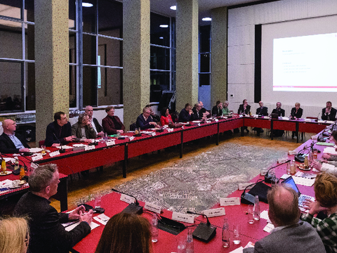 Foto, das Menschen zeigt, die an einem U-förmigen Tisch sitzen und beraten, im Hintergrund wird eine Präsentation an die Wand projiziert.