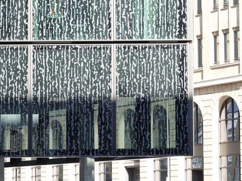Die Glasfassaden des Axel-Springer-Neubaus sind teils vogelfreundlich gestaltet. Ein hellgraues Muster, das an ein Bambus-Dickicht erinnert, ist vollflächig auf das grünlich getönte Glas gedruckt. Eine spitze Ecke der Fassade, die auf schlanken Pfeilern zu schweben scheint, ragt weit ins Bild und nimmt Dreiviertel des Fotos ein. Ein breiter Schattenstreifen verdunkelt den unteren Bereich. Im Hintergrund die sonnenbeschienene Fassade des denkmalgeschützten Krausenhofes mit hohen Arkadenfenstern.