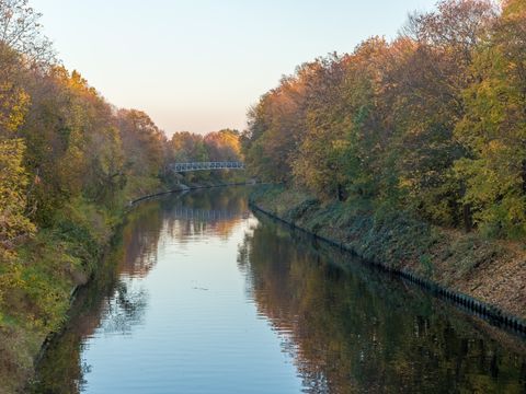 Das Foto zeigt einen etwas erhöhten Blick auf den Teltowkanal. Herbstlich gefärbte Bäume säumen ihn zu beiden Seiten. Etwas entfernt überspannt eine Brücke das ruhige Gewässer. An dieser Stelle macht der Wasserlauf eine langgezogene Biegung nach rechts. Verschwommen reflektiert die leicht gekräuselte Wasseroberfläche die dichte Ufervegetation in Gelb-, Rot- und Grüntönen sowie den blassblauen Himmel.