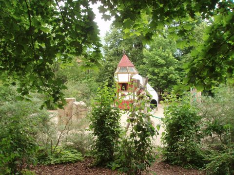 Der Spielplatz der Reinhardswald-Grundschule im Bezirk Friedrichshain-Kreuzberg ist von grünen Büschen, Bäumen und Sträuchern umgeben. Inmitten einer Sandfläche ragt ein Kletterturm mit rotgestrichener Spitze in die Höhe. Eine Röhrenrutsche aus Edelstahl windet sich von dem eckigen Spielhaus zu Boden. Links stehen zwei Natursteinblöcke im hellgelben Pudersand.
