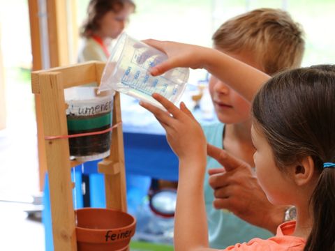 Beim Workshop „Boden schätzen“ im HELLEUM gießt ein Mädchen mit braunen Seitenzöpfen Wasser in eine selbst gebaute Filterstation. Ein blonder Junge daneben beobachtet, wie Wasser aus einem durchsichtigen Gefäß mit Erde in einen Blumentopf tropft. Mit einem Gummiband ist das Gefäß in einer leiterartigen Holzkonstruktion befestigt. Die Hand einer erwachsenen Person außerhalb des Bildes deutet darauf.