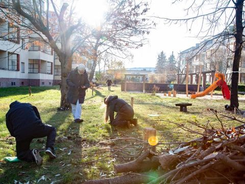 Ein Blick in einen umgestalteten Wohnhof der Genossenschaft „Märkische Scholle“ in Tempelhof. Ein Mann und eine Frau in dicken Jacken knien im Gras auf dem weiten Streifen zwischen zwei Wohnblocks. Mit Blick zum Boden steht eine zweite Frau vor ihnen. Gleißendes Sonnenlicht scheint durch die Krone eines Baums mit wenigen braunen Blättern. In der rechten unteren Bildecke sind Zweige und Äste zu einem kleinen Haufen gestapelt. Dahinter ein kleiner Spielplatz.