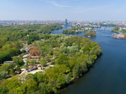 Aus großer Höhe ein Blick auf den ausgedehnten grünen Plänterwald, der sich links im Bild parallel zur tiefblauen Spree erstreckt und in den Treptower Park über-geht. Im Vordergrund ragt zwischen Bäumen das rote Riesenrad des ehemaligen Spreeparks auf. In weiter Ferne lassen sich auf der Spree die Halbinsel Stralau und die Rummelsburger Bucht erkennen. Das markante Hochhaus der Treptowers zeichnet sich vor dem blauen Himmel ab.