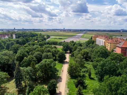 Ein Blick aus großer Höhe auf einen schnurgeraden Weg, der von Wiesen und satt grünen Bäumen gesäumt wird. Darüber spannt sich blauer Himmel mit weißen Wolken. Etwas zurückgesetzt reihen sich rechterhand Altbauten aneinander. Der Weg führt auf den ehemaligen Flugplatz Tempelhof, heute als Tempelhofer Feld bekannt, zu. Auf der riesigen Fleifläche sind die früheren Start- und Landebahnen von Wiesen umgeben.