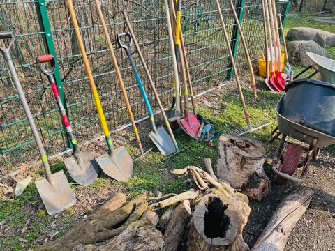 Im Sonnenschein lehnen an einem grünen Zaun verschiedene Gartengeräte wie Spaten, Schaufeln oder Harken. Die Griffe sind zum Teil mit bunten Bändern umwickelt. Auf einem Weg davor liegen am Rand helle Totholz Stücke neben leeren Schubkarren.