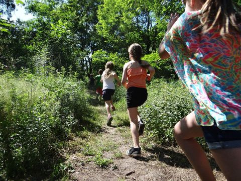 Eine Gruppe Kinder rennt hintereinander einen schmalen sonnenbeschienenen Pfad entlang. Zu beiden Seiten wachsen hohe Pflanzen. Der ausgetretene Pfad führt in einen schattigen Waldbereich. Vorneweg laufen zwei Jungen, ihnen folgen drei Mädchen etwa 10 Jahre alt. Alle tragen Sommerkleidung.