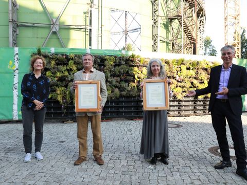 Ein Gruppenfoto vor dem historischen Wasserturm des Euref-Campus. Von links nach rechts stehen Anette Nawrath (Vorstandsvorsitzende Stiftung Naturschutz Berlin), Jens Scharon (Preisträger), Gülcan Nitsch (Preisträgerin) und Stefan Tidow (Staatssekretär für Umwelt und Klimaschutz) mit Abstand nebeneinander. Herr Scharon und Frau Nitsch halten die holzge-rahmten Urkunden hoch.