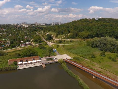 Das Umweltbildungszentrum Kienbergpark von oben. Das langgestreckte Gebäude mit Holzfassade und Flachdach befindet sich auf einer auf 60 Meter langen Holz-Plattform. Vor dem Gebäude stehen Tische und Stühle unter Sonnenschirmen. Rechts davon führt der Wuhlesteg, eine 300 Meter lange Brücke, auf die Plattform. Hinter dem Umweltbildungszentrum erstreckt sich links eine Kleingartenanlage, rechts erhebt sich der dicht bewaldete Kienberg, dessen Gipfel mit einer Seilbahn erreichbar ist.