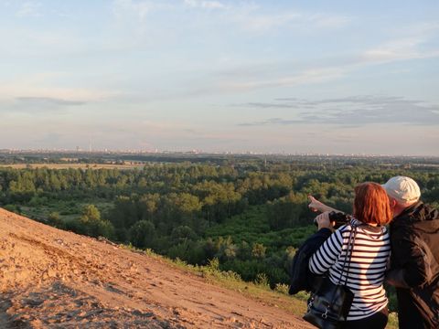 In der Abendsonne stehen am rechten unteren Bildrand abgewandt eine ältere Frau und ein Mann auf einem Sandweg. Sie schauen von einem Hügel auf eine Landschaft mit Laubwäldern und Wiesen. Am Horizont zeichnet sich die Silhouette von Berlin ab. Der blaue Himmel wird von einigen Wolkenbändern durchzogen. Die Frau hat halblanges, rotes Haar und trägt ein schwarz-weiß geringeltes Shirt. Sie deutet in die Ferne und hält ein Fernglas. Der Mann trägt ein beiges Käppi und eine schwarze Jacke.