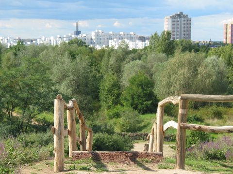 Das weitläufige Gelände des Freizeitparks Marienfelde im Sonnenschein. Eine breite Treppe mit beidseitigem Holzgeländer führt abwärts in eine grüne Senke. Ein schmaler Sandweg verläuft nach links und wird von Wiesen mit Büschen und üppigen Laubbäumen gesäumt. In weiter Ferne erheben sich hinter dem Park dicht an dicht gebaute Hochhäuser in den blauen Himmel.