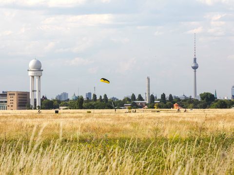 Ein Blick aus der Ferne über das Tempelhofer Feld. Auf den weiten gelblichen Wiesenflächen sind als winzige Punkte Menschen auszumachen. Einige sind mit dem Fahrrad unterwegs. Ein Gleitschirm schwebt in der Luft. Am linken Bildrand ist das ehemalige Flughafengebäude angeschnitten, daneben steht der weiße frühere Radarturm. Im Hintergrund ragen hinter Bäumen Häuser sowie der Fernsehturm in den bewölkten Himmel.