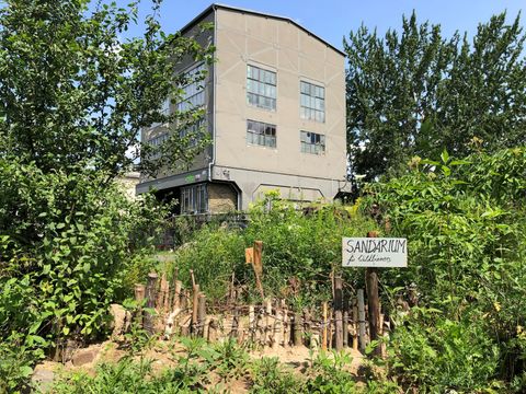 Das Foto zeigt einen wild bewachsenen Gartenbereich mit dichtem Grün, im Vordergrund ein mit entsprechendem Schild gekennzeichnetes "Sandarium für Wildbienen", umgeben von Holzstäben. Im Hintergrund steht ein mehrstöckiges, graues ehem. Bahngebäude mit großen Fenstern, flankiert von hohen Bäumen. Die Stadtnatur-Szenerie wirkt naturnah, biodivers und urban, bei sonnigem Wetter unter blauem Himmel.